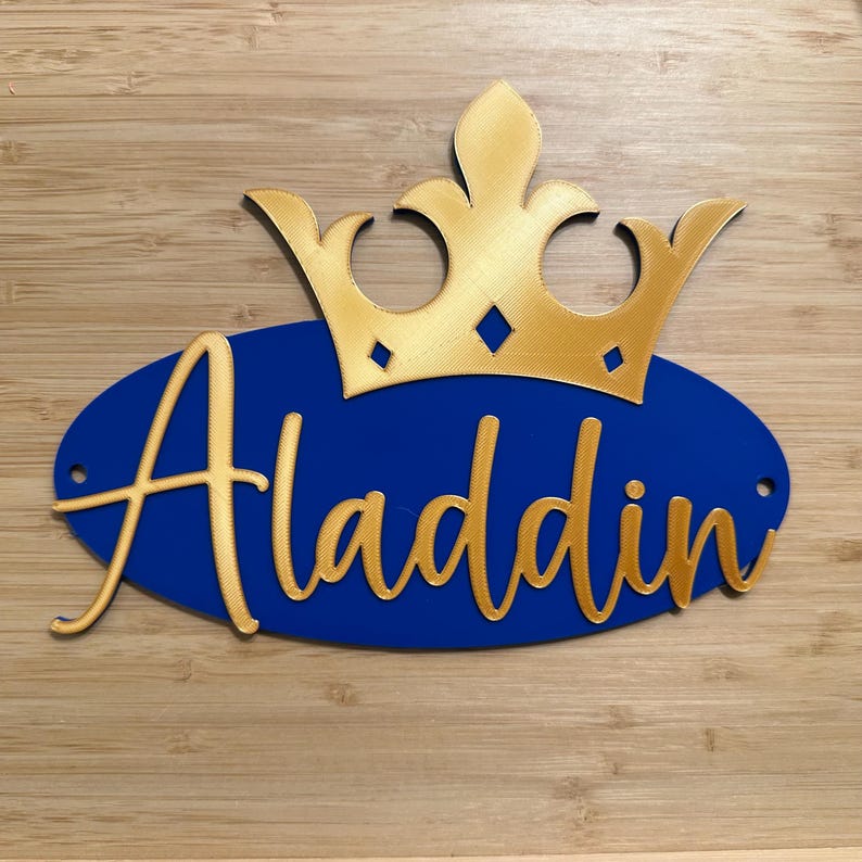 May include: A gold-colored crown sits atop a blue oval with the name "Aladdin" in gold script. The design is set against a light brown wooden background. The crown has diamond-shaped cutouts.