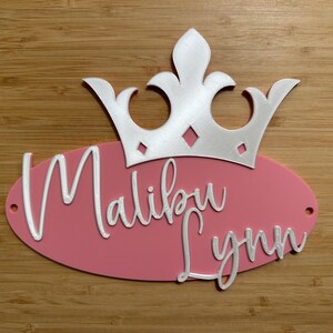 May include: A pink oval sign with the name "Malibu Lynn" in white script, topped with a white crown. The sign has two small holes on the right side.