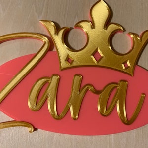 May include: A custom name sign featuring the name "Zara" in gold script, set against a pink oval background. A gold crown sits above the name. The sign has two small holes for hanging.