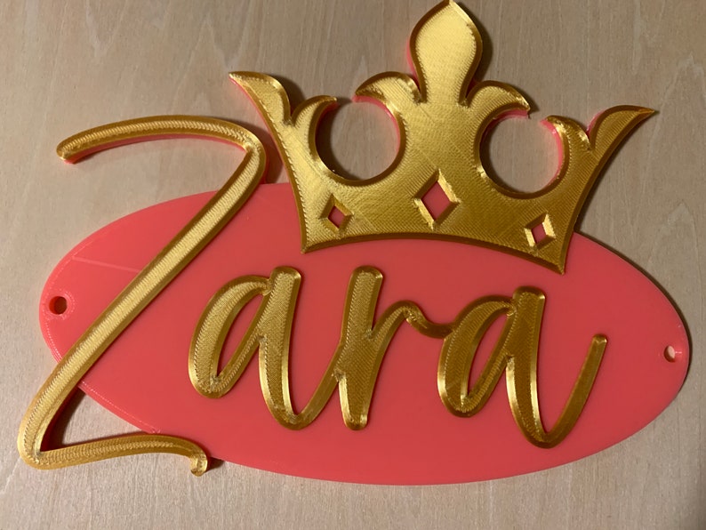 May include: A custom name sign featuring the name "Zara" in gold script, set against a pink oval background. A gold crown sits above the name. The sign has two small holes for hanging.