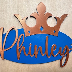 May include: A blue oval sign with the name "Phinley" in copper-colored script, topped with a copper crown. The sign has two small holes for hanging and is set against a light wood background.
