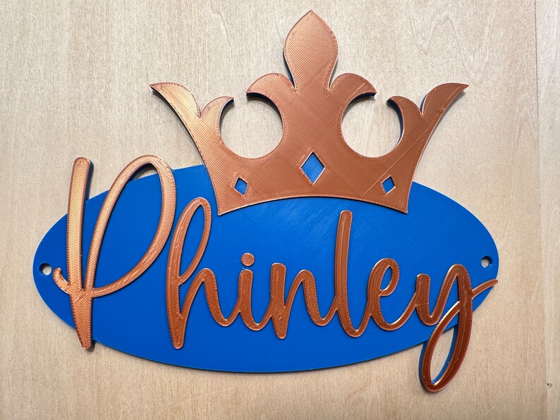 May include: A blue oval sign with the name "Phinley" in copper-colored script, topped with a copper crown. The sign has two small holes for hanging and is set against a light wood background.