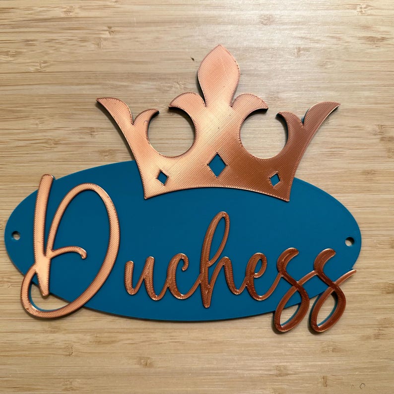 May include: A teal oval sign with the word "Duchess" in copper-colored script, topped with a copper-colored crown. The sign has two small holes for hanging and is set against a wooden background.