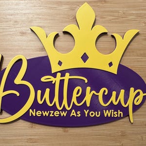 May include: A yellow crown sits atop a purple oval with the name "Buttercup" in yellow script. Below the name is the phrase "Newzew As You Wish" in yellow. The sign is on a light brown wooden surface.
