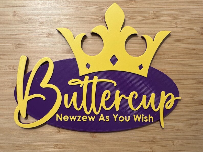 May include: A yellow crown sits atop a purple oval with the name "Buttercup" in yellow script. Below the name is the phrase "Newzew As You Wish" in yellow. The sign is on a light brown wooden surface.