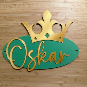 May include: A personalized sign featuring the name "Oskar" in gold script on a teal oval, topped with a gold crown. The sign is designed with a 3D effect and includes two small holes for hanging.
