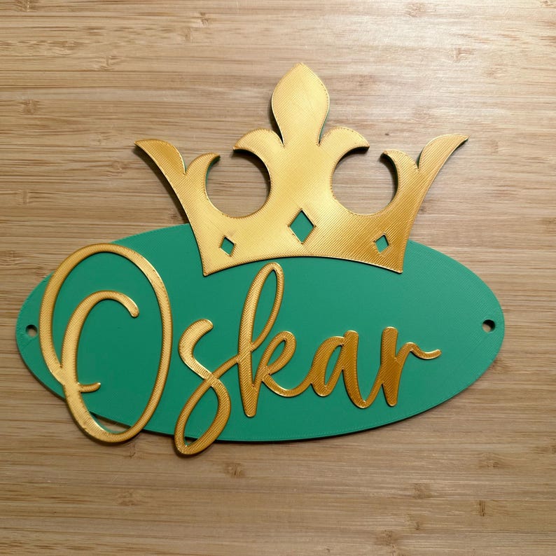 May include: A personalized sign featuring the name "Oskar" in gold script on a teal oval, topped with a gold crown. The sign is designed with a 3D effect and includes two small holes for hanging.
