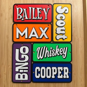 May include: A collection of colorful license plates with names in bold white lettering. Names include "Bailey", "Max", "Bingo", "Whiskey", "Cooper", and "Scout". Each plate has a different background color, including red, orange, yellow, green, blue, and purple.