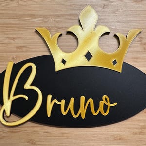 May include: A decorative sign featuring a gold crown above a black oval. The name "Bruno" is written in gold script on the oval. The sign has two small holes for hanging and is set against a wooden background.