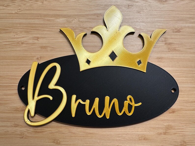 May include: A decorative sign featuring a gold crown above a black oval. The name "Bruno" is written in gold script on the oval. The sign has two small holes for hanging and is set against a wooden background.