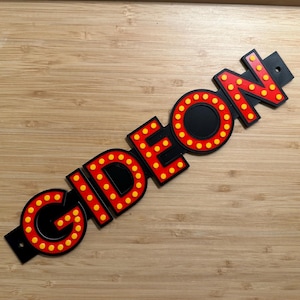 Puede incluir: Letrero negro con el nombre "GIDEON" en letras rojas con luces de puntos amarillos. El letrero está inclinado sobre una superficie de madera. Las letras están delineadas en rojo con luces de puntos amarillos.