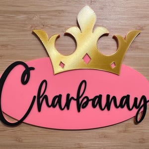 May include: A pink oval-shaped sign with the name "Charbanay" in black cursive script, topped with a gold-colored crown. The sign is set against a wooden background.