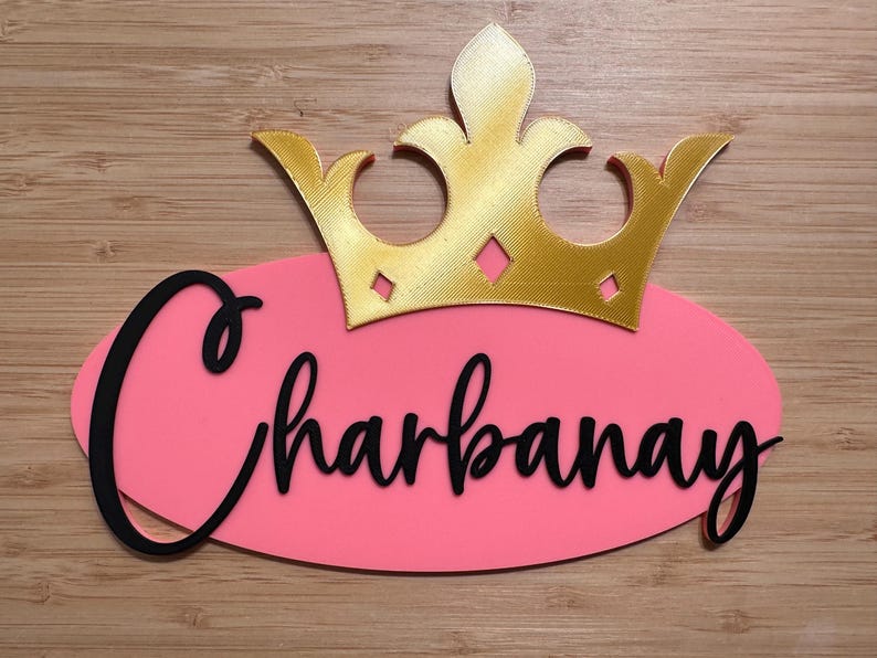 May include: A pink oval-shaped sign with the name "Charbanay" in black cursive script, topped with a gold-colored crown. The sign is set against a wooden background.