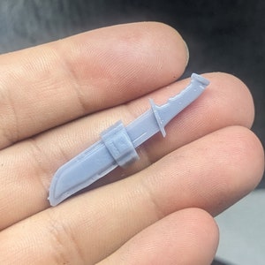 Puede incluir: Un modelo de cuchillo de plástico gris con una funda. El cuchillo tiene una hoja dentada y un mango negro.