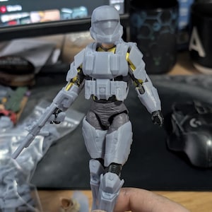 May include: A gray plastic action figure of a soldier wearing a helmet and armor. The figure is holding a gun in one hand and is standing on a table.