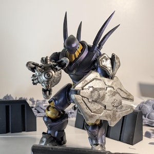 May include: A dark blue and silver action figure of a robot with a weapon in one hand and a shield in the other. The robot has a yellow face and sharp spikes on its back.