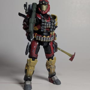 May include: A red and yellow action figure of a soldier wearing a helmet with a visor and holding a green weapon. The figure is wearing a red and black suit with a yellow belt and has a black axe on its back.