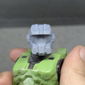 Könnte beinhalten: Nahaufnahme einer grünen und grauen Actionfigur. Die Figur hat einen grauen Helm und eine grüne Rüstung. Die Figur wird von einer Hand gehalten.