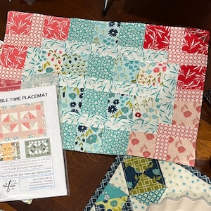 May include: Two colorful placemats with patchwork designs. One features a pink and white geometric pattern, the other has a mix of floral and geometric patterns in shades of pink, teal, and red. A package with the text "DOUBLE TIME PLACEMAT" is also visible.