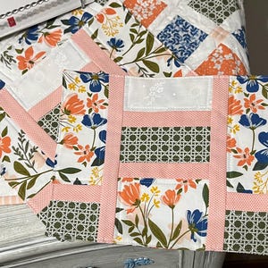 May include: Floral placemats with a white background, featuring orange and blue flowers, green leaves, and a pink border. The placemats have a rectangular shape and a decorative woven pattern.