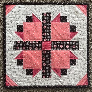 May include: A square quilt with a central floral design in pink, black, and white. The quilt features a repeating pattern of small flowers and geometric shapes. The border is a dark brown with a repeating pattern. The quilt is approximately 12 inches square.