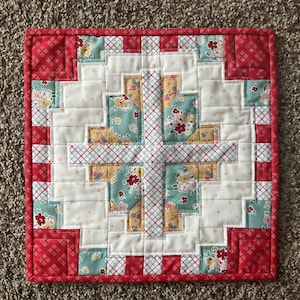 May include: A square, handmade quilt with a patchwork design. The quilt features a central cross pattern in white, yellow, and floral fabrics, surrounded by red and white patterned borders. The quilt is likely used as a decorative item.