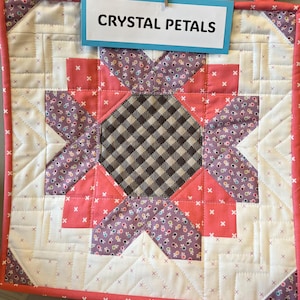 May include: A handmade quilt with a starburst design. The quilt features a central square of brown and white checkered fabric, surrounded by pink, purple, and white patterned fabric. The text "CRYSTAL PETALS" is displayed on a blue sign.
