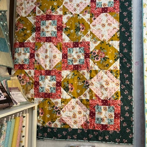 May include: A large, colorful quilt with a floral pattern, displayed on a wall. The quilt features squares of pink, white, and mustard yellow fabric, with a dark green border. The quilt is a patchwork design, with a variety of floral patterns.
