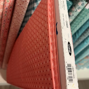 May include: A stack of coral-colored fabric with a polka dot pattern. The fabric is labeled "Bee Basics" and "C6405" with the color "Coral". The fabric is 43/44 inches wide and made of 100% cotton. Other fabric rolls are visible in the background.