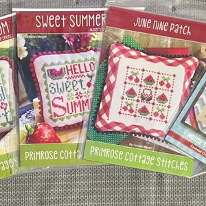 May include: A collection of cross-stitch pattern packets. The packets feature designs with text like "Plant, Grow, Bloom" and "Sweet Summer." The color palette includes red, green, and blue, with images of fruit and floral motifs. The packets are labeled "Primrose Cottage Stitches."