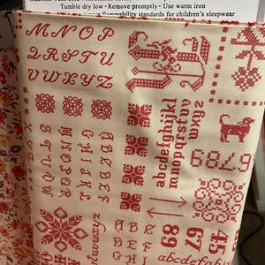 May include: Cream-colored fabric with a red cross-stitch alphabet and numbers. The fabric features various cross-stitch patterns, including floral and geometric designs. The text "For reorder call (888)768-8454" is visible.