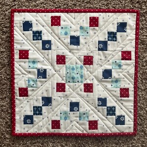May include: A square, handmade quilt with a red border. The quilt features a repeating pattern of red, white, and blue squares, with a white background and small star details. The quilt is approximately 24 inches square.