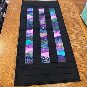 May include: A rectangular black table runner with three vertical columns of colorful, patterned fabric. The columns feature a mix of purple, pink, and teal fabrics. The runner is quilted with horizontal lines.