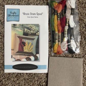 May include: An assortment of cross-stitch supplies, including a pattern booklet titled "Illinois State Spool" by Crafty Bluebonnet Designs. The image also shows a package of embroidery floss in various colors and a piece of tan Aida cloth.