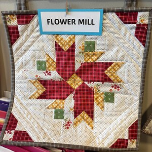 May include: A quilted wall hanging with a central starburst design. The star is composed of red plaid and yellow floral fabrics, with green square accents. The background is a white fabric with a small dotted pattern. The text "FLOWER MILL" is displayed on a blue sign.