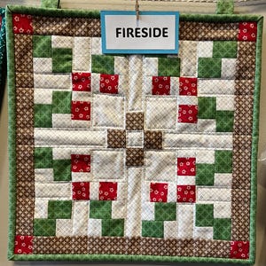 May include: A square quilted wall hanging with a patchwork design. The quilt features a repeating pattern of green, white, red, and brown squares. The word "FIRESIDE" is displayed on a blue sign attached to the top.