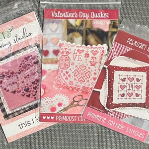 May include: Three cross-stitch pattern packets in plastic sleeves. The packets feature Valentine's Day themes with hearts and the text "Valentine's Day Quaker", "February Nine Patch", and "Sweet Wing Studio". The color scheme is primarily pink and red.