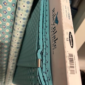 May include: A stack of turquoise fabric with a small dot pattern, held together with a silver clip. The fabric is labeled "Riley Blake Designs" and "Bee Basics". The fabric is 43/44" wide and 100% cotton. The pattern is C6405.