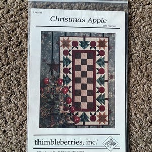 May include: A clear plastic bag containing a pattern for a Christmas Apple table runner. The pattern features a quilt design with red apples, green trees, and star accents. The text "Christmas Apple" is visible.