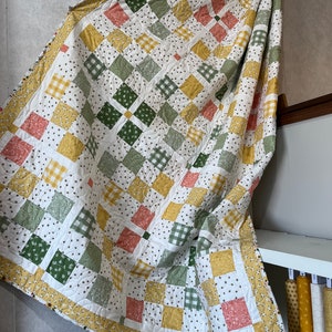 May include: A handmade quilt with a patchwork design. The quilt features a white background with squares in shades of green, yellow, and peach. Some squares have floral or gingham patterns. The border is yellow with a floral pattern. The quilt is draped over a surface.
