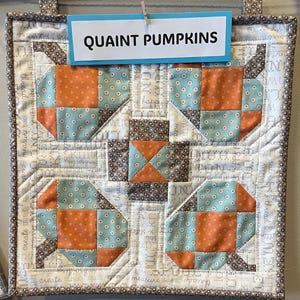 May include: A quilted wall hanging with a pumpkin design. The pumpkins are made of orange and teal fabric squares, set against a white background with text. The sign at the top reads "QUAINT PUMPKINS".
