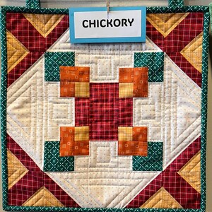 May include: A quilted wall hanging with a patchwork design. The quilt features a central square with red, orange, and white patterned squares. The word "CHICKORY" is displayed on a blue sign. The border is green.