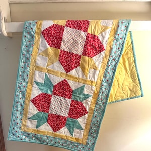 May include: A handmade quilt with a star pattern. The quilt features red, yellow, white, and teal fabrics. The star blocks are framed by yellow and teal borders. The quilt is hanging on a white shelf.