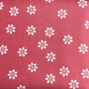 May include: Close-up of a red fabric with a repeating pattern of white and pink daisy-like flowers. The flowers are evenly spaced across the surface, creating a cheerful and delicate design. The fabric has a slightly textured appearance.