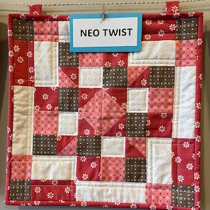 May include: A square quilted wall hanging with a patchwork design. The quilt features red, white, and brown fabrics with floral and polka dot patterns. The text "NEO TWIST" is on a blue tag attached to the top.