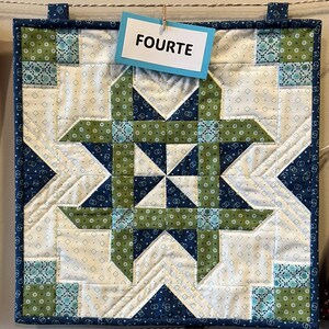 May include: A square quilted wall hanging with a starburst design in white, navy blue, and green. The word "FOURTE" is printed on a small tag hanging from the top. The edges are finished with a navy blue border.