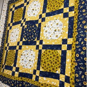 Puede incluir: Un edredón grande, hecho a mano, con un tema de girasoles. El edredón tiene un diseño de patchwork con cuadrados alternos de girasoles amarillos, patrones florales blancos y formas geométricas azul marino. El borde es azul marino con detalles de girasoles.