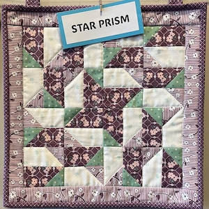 May include: A handmade quilt with a star pattern, featuring squares of white, green, and purple floral fabric. The quilt is framed with a floral border and the text "STAR PRISM" is displayed on a blue tag.
