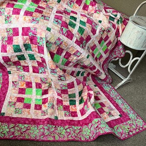 May include: A handmade quilt with a vibrant patchwork design. The quilt features squares of pink, green, and peach fabrics, creating a checkerboard pattern. The border is a darker pink with a floral design. The quilt is draped over a surface.