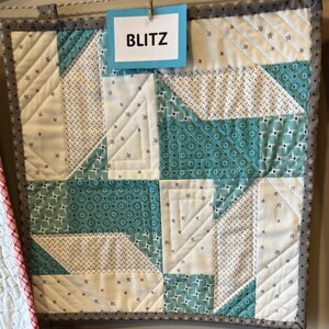 May include: A quilted wall hanging with a geometric pattern in white, teal, and patterned fabrics. The word "BLITZ" is printed on a blue tag attached to the top. The quilt has a gray border.